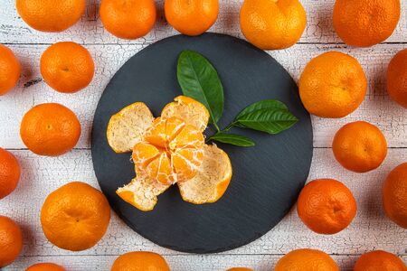 One opened tangerine with leaves on round black plate surrounded by whole bright orange tangerines on cracked white wooden table. Fruits have natural imperfections. Healthy eating and dieting concept.の写真素材