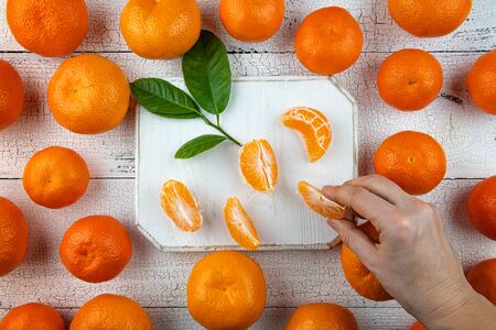 Human hand is taking tangerine segments from square white cutting board surrounded by whole orange...