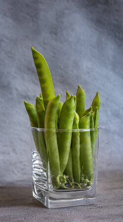 Whole green sweet pea pods in drinking glass. One pod sticks out of the pile and seeds are visible through transparent pod. Vertical gray background with copy space. Healthy eating and dieting conceptの写真素材
