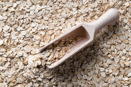 Wooden scoop with few flakes of dry rolled oats in it. Scoop is on layer of raw rolled oats. Source...