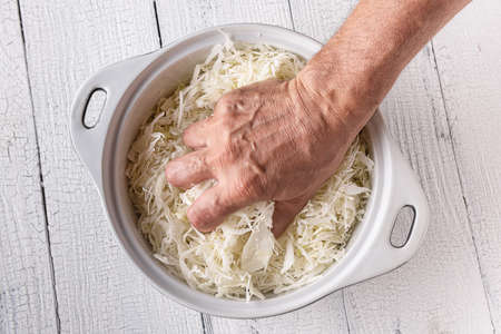 Hand of man squeezing grated cabbage in ceramic pan on old wooden white table surface made of planks. Cooking stage for coleslaw, fermented cabbage and salad. Background for healthy food concept.の写真素材