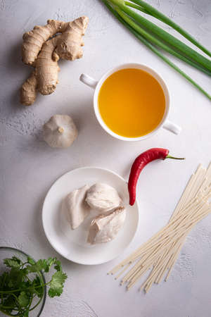 Spicy asian soup ingredients. Ginger, garlic, chicken bouillon and meat, red chilli pepper, cilantro and scallion, japanese noodles. White textured stone background. Flat lay. Healthy food concept.の写真素材