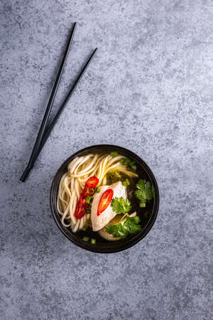 Spicy asian soup. Chicken bouillon and meat, red chilli pepper, fresh cilantro and scallions, japanese noodles in black soup bowl with chopsticks on gray surface. Vertical orientation. Copy space.の写真素材