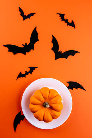 Whole raw orange pumpkin on flying white round plate with black wings and hand carved black bat silhouettes on vertical orange paper background. Flat lay. Funny image with Halloween symbols.の写真素材