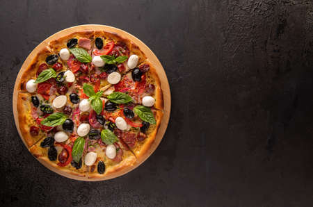 Italian pizza with ham, salami, black olives, mozzarella cheese, red tomatoes and basil leaves on...
