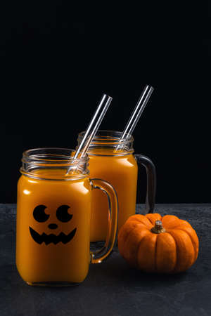 Two square glass jars with non-alcoholic pumpkin mocktail and glass straws on black textured surface. Jar is decorated with black halloween face. Vertical background with copy space. Selective focus.の写真素材