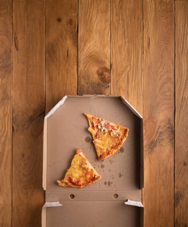Partly visible used opened pizza box with two ugly pieces of cold pizza, stains and crumbs inside on natural wooden background. Vertical orientation. Copy space. Home delivery and stay home concept.の写真素材
