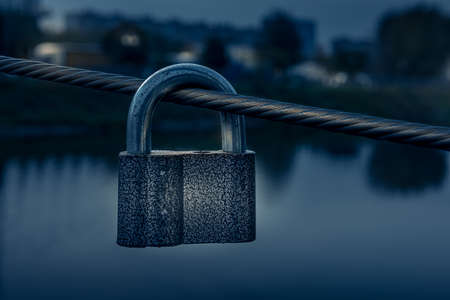 Metal padlock hanging on steel cable of bridge fence in dusk. Unfocused dark cityscape with...