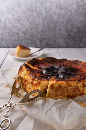 Cut homemade basque burnt cheesecake in baking paper and silver dessert tongs on linen napkin and on white wooden table. Selective focus. Spanish dessert from San Sebastian city of Spain. Copy space.の写真素材