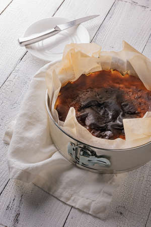 Homemade basque burnt cheesecake in baking tin on white linen napkin and on textured wooden table. Knife on plate at background. Selective focus. Spanish dessert from San Sebastian city of Spain.の写真素材