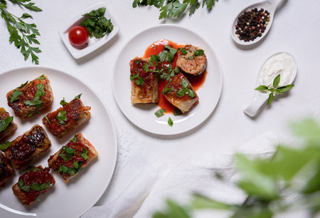 Flat lay with stuffed cabbage rolls in tomato sauce, parsley and few ingredients on white plates, including sour cream and pepper. Unfocused parsley at foreground. White tesxtured stone background.の写真素材