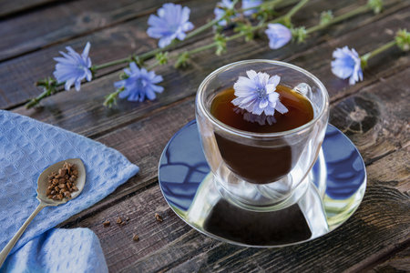 Drink made of chicory roots in thermo glass cup, blooming chicory plants, blue linen napkin and spoon with granulated chicory root powder on brown wooden table. Caffeine free alternative to coffee.の写真素材