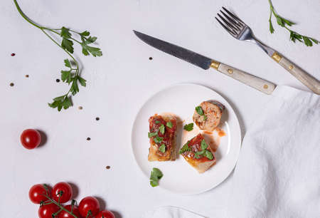 Cabbage rolls in tomato sauce, few ingredients on white plates, sour cream and pepper and white linen napkin. One roll is cut. White textured stone background. Flat lay. Horizontal orientation.の写真素材