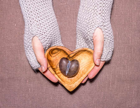 Wooden heart shaped plate with ripe malformed imperfect plum in hands of caucasian woman with nude manicure in knitted warm fingerless gloves or mittens. Lilac linen background. Directly above view.の写真素材