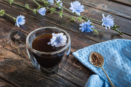 Caffeine free alternative to coffee drink made of chicory in thermo glass cup, blooming chicory stems, blue linen napkin and spoon with granulated chicory root powder on brown natural wooden table. .の写真素材