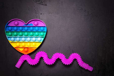 Flat lay with popping toy rainbow heart and bent as wave pink pop tube toy. Plastic antistress multicolored children toy set on black textured concrete background. Large copy space. Horizontal orientation.の写真素材