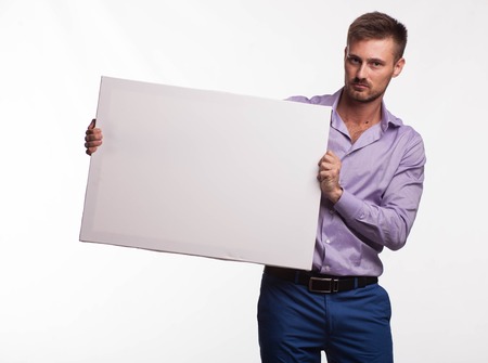 Young serious man portrait of a confident businessman showing presentation, pointing paper placard gray background. Ideal for banners, registration forms, presentation, landings, presenting concept.の写真素材