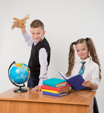 Happy boy and girl sit at a school desk, isolated on white background. Boy plays by airplane. Girl is reading a book. Ideal for banners, registration forms, presentation, landings, presenting concept.の写真素材