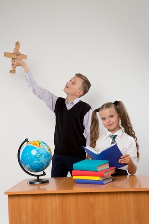 Happy boy and girl standing at a school desk, isolated on white background. Boy plays by airplane. Girl is reading a book. Ideal for banners, registration forms, presentation.の写真素材