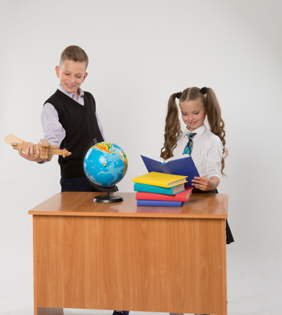 Happy boy and girl sit at a school desk, isolated on white background. Boy plays by airplane. Girl is reading a book. Ideal for banners, registration forms, presentation, landings, presenting concept.の写真素材
