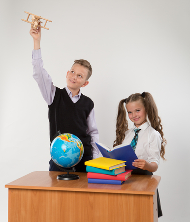Happy boy and girl sit at a school desk, isolated on white background. Boy plays by airplane. Girl is reading a book. Ideal for banners, registration forms, presentation, landings, presenting concept.の写真素材