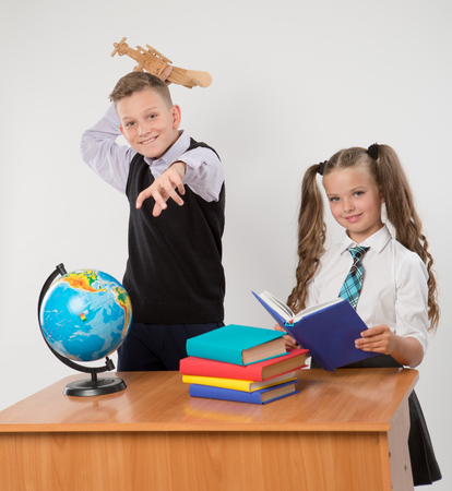 Happy boy and girl sit at a school desk, isolated on white background. Boy plays by airplane. Girl is reading a book. Ideal for banners, registration forms, presentation, landings, presenting concept.の写真素材