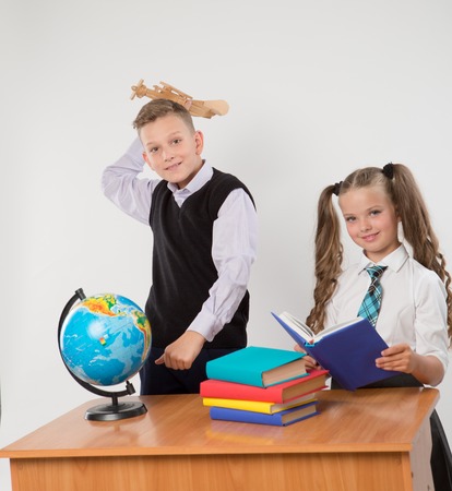 Happy boy and girl sit at a school desk, isolated on white background. Boy plays by airplane. Girl is reading a book. Ideal for banners, registration forms, presentation, landings, presenting concept.の写真素材