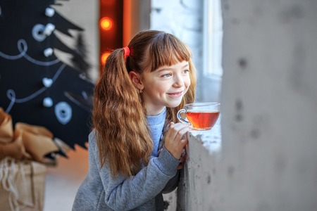  A little girl dreams and looks at the window. The child drinks tea. Concept Happy Christmas, New Year, holiday, winter, childhood.の写真素材