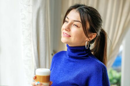 A happy woman stands by the window and drinks a lot. The concept of lifestyle, comfort, success.の写真素材
