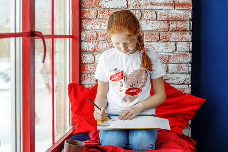 Red-haired child sits on the window sill and paints. The concept is childhood, learning, drawing, hobbies, education, talent.の写真素材