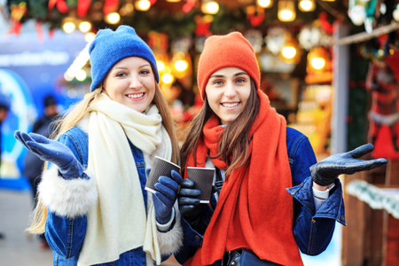 two young girls in red and blue hats and scarves walk around the Christmas market, drink coffee,...