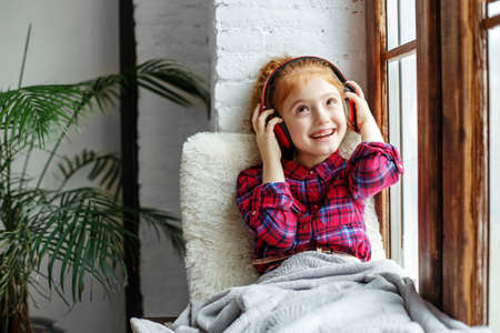 A teenage girl with red curly hair enjoys listening to music sitting on the windowsill. Listen to music school girlの写真素材