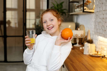 Young girl with red hair, teenager, in a modern kitchen playing with an orange, drinking orange juice, happy.の写真素材
