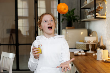 Young girl with red hair, teenager, in a modern kitchen playing with an orange, drinking orange...