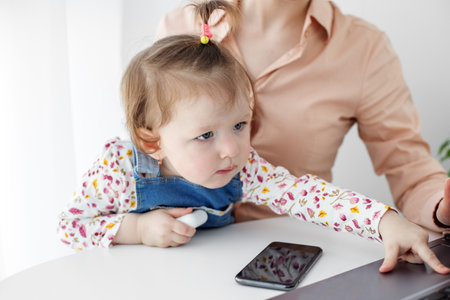 A small kid presses the keys of a laptop. The little daughter prevents her mother from working at the computer. Work from home online. Work in the decree.の写真素材