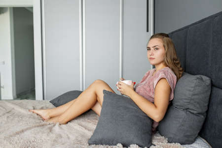Teenage girl in gentle pink pajamas. Drink delicious coffee in bed. Young woman resting at home.