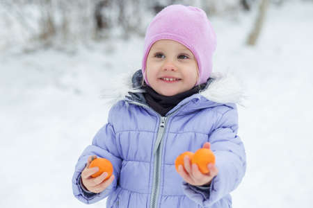 A little girl walks in winter clothes in a snow-covered park. Delicious tangerines.の写真素材