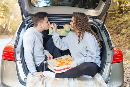 A man and a woman are eating pizza near the car. Picnic in the fall.