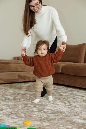 Adorable baby holding mother's hands, little daughter walking through living room. The concept of...