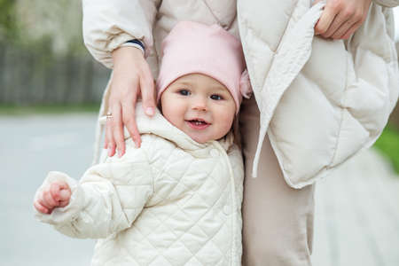 Little baby girl learns to walk with mom's help outside. Young mother and daughter toddler walk in...