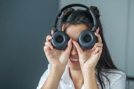Beautiful young adult brunette girl holds headphones, makes grimace and smile. Concept of music, study, audio books and workの写真素材