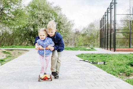 Child boy teaches little sister how to ride a children's scooter. Active rest on playground in spring day.の写真素材