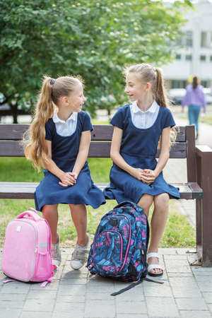 Two children of schoolgirls sit on bench near school, have fun and communicate. Concept of back to school, learning, friendship and childhoodの写真素材