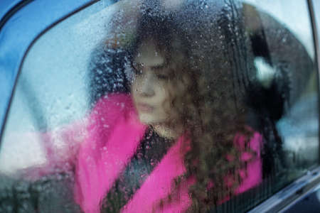 Sad woman looks out foggy car window. Window with rain drops. Curly-haired woman is riding in car in...