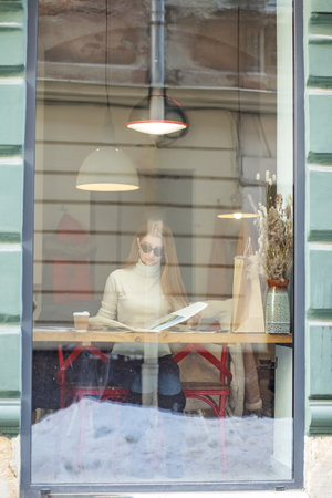 Young adult woman is reading magazine in cafe and drinking hot drink. Girl behind glass in cafe