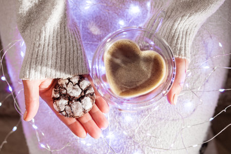 Female hands holding cup of coffee and cookies in Christmas lights. Heart shape.の写真素材