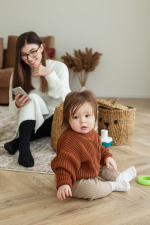 Mom and baby toddler in room. Girl is playing. Mother is using smartphone