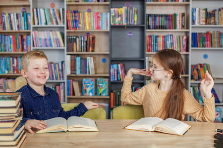 Little school children reading books together while sitting at table in library. Boy and girl study...