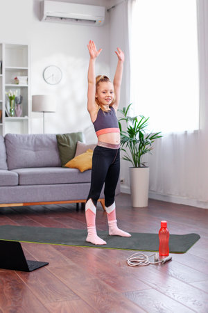Reusable water bottle. Distant training with personal trainer. Child girl in sportswear watching online video on laptop and doing fitness exercises at home.の写真素材
