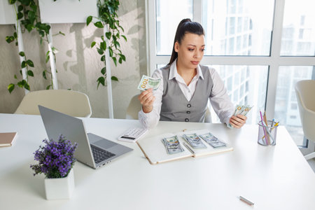 Sad business woman sits at work desk in front of laptop and counts cash. Concept of low profits, low salary and financial problems.の写真素材